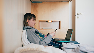 Tonårstjej sitter med dator och telefon i en säng.